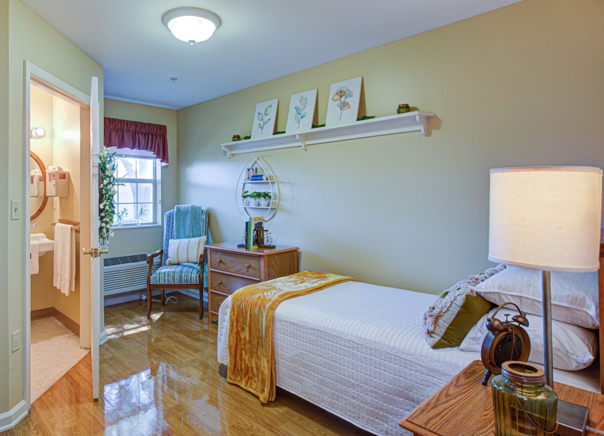 A cozy bedroom with a single bed, adorned with decorative pillows and a throw blanket. The room features wooden flooring, a nightstand, an armchair by the window, and a shelf with framed pictures.