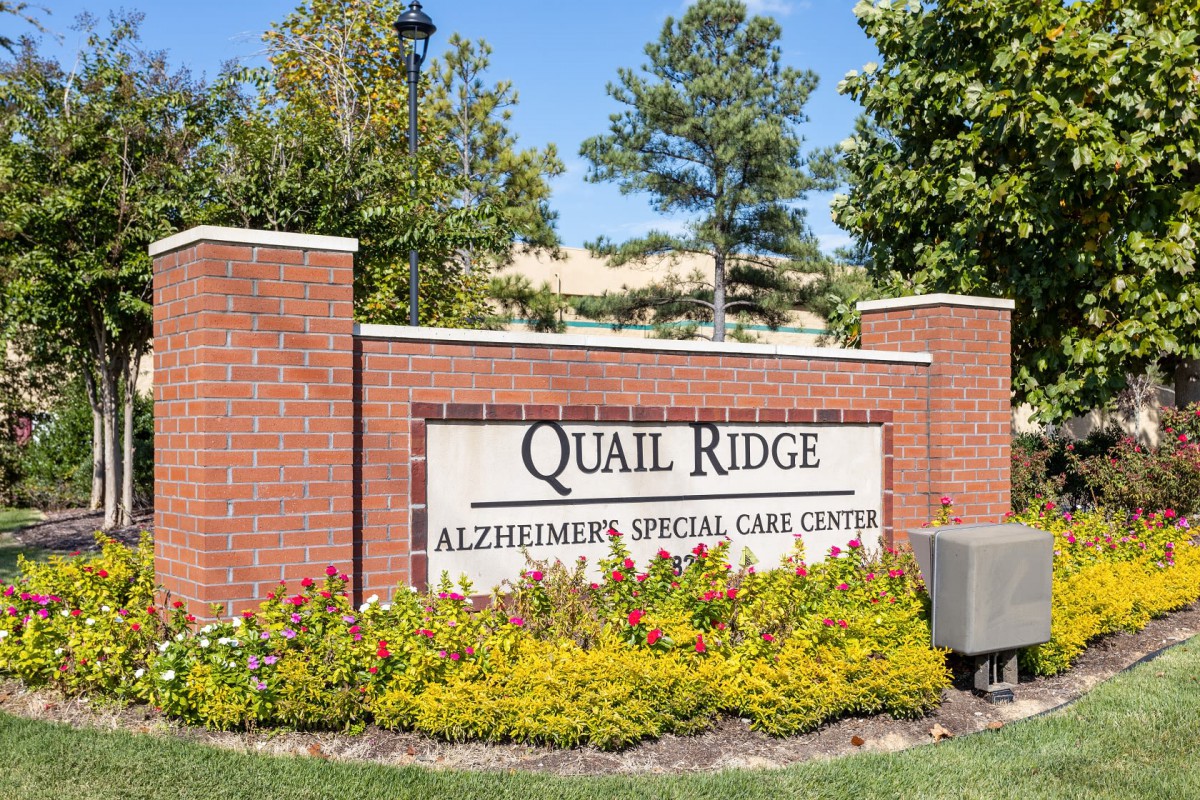 Sign for Quail Ridge Alzheimer's Special Care Center set against a brick wall with surrounding greenery and trees.