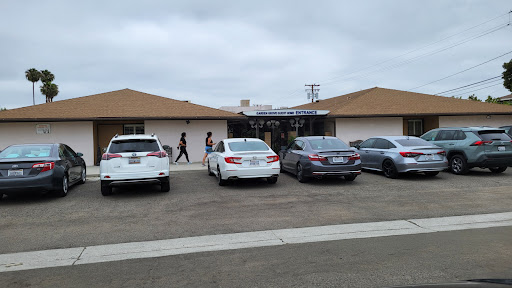 A view of a parking lot in front of a building with several cars and people walking by.