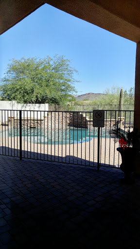 A serene view of a swimming pool surrounded by a fence, with a clear blue sky and lush greenery.