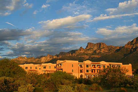A warm sunset casts a golden hue over a desert landscape with mountains in the background and a building in the foreground.