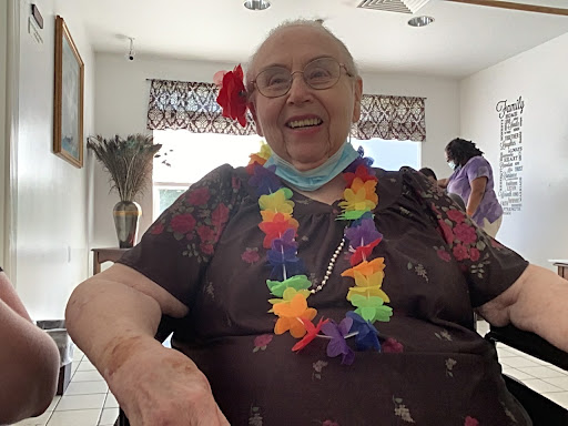 An elderly woman with a joyful smile, adorned in a vibrant Hawaiian-style lei and a flower in her hair, enjoying a cheerful gathering indoors.
