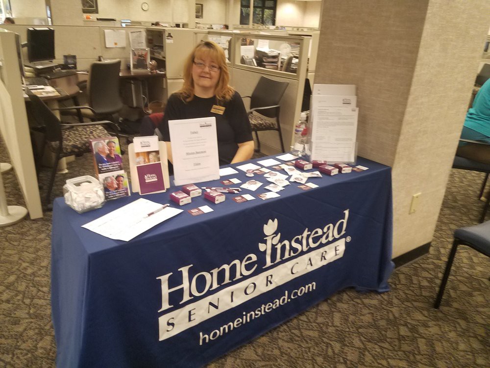 A representative sits at an informational booth for Home Instead Senior Care, showcasing promotional materials and brochures.