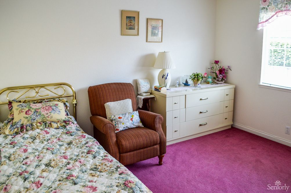 A cozy bedroom with a floral bedspread, an armchair, and a dresser on a pink carpet.