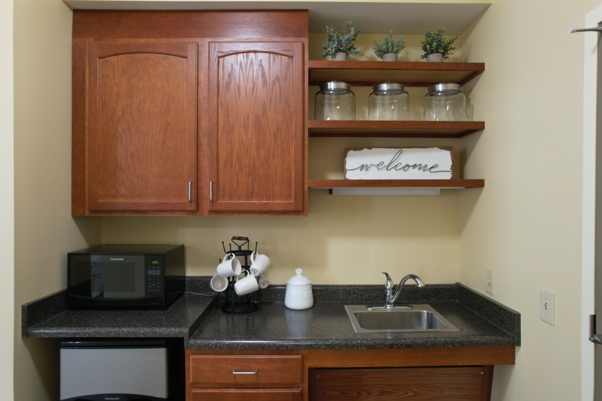 A cozy kitchen corner featuring wooden cabinets, open shelves with decorative plants and jars, a microwave, and a small sink.