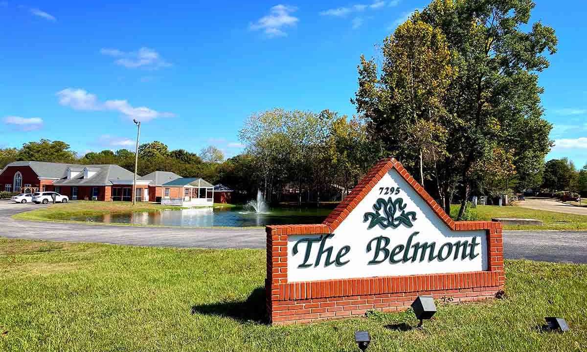 A scenic view of a brick sign for 'The Belmont' with a pond and fountain in the background.