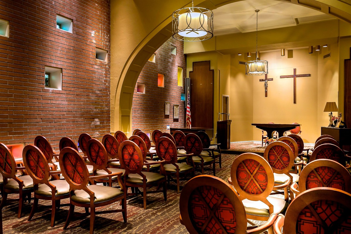 An interior view of a chapel with arranged seating and a cross on the wall.