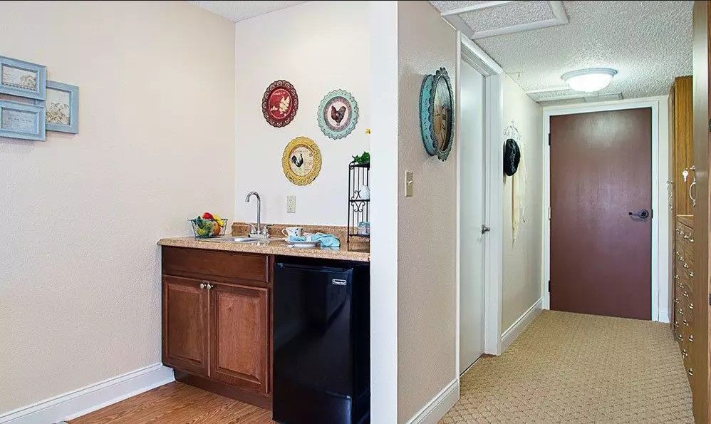 A cozy kitchenette area with decorative plates on the wall, featuring a mini fridge, sink, and wooden cabinets, adjacent to a hallway.