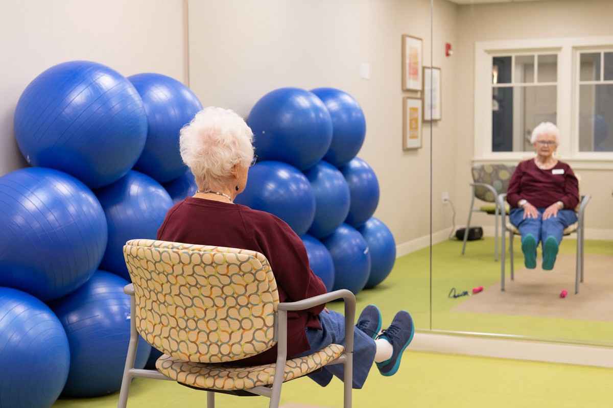 An elderly person sitting on a chair in front of a mirror, surrounded by blue exercise balls.