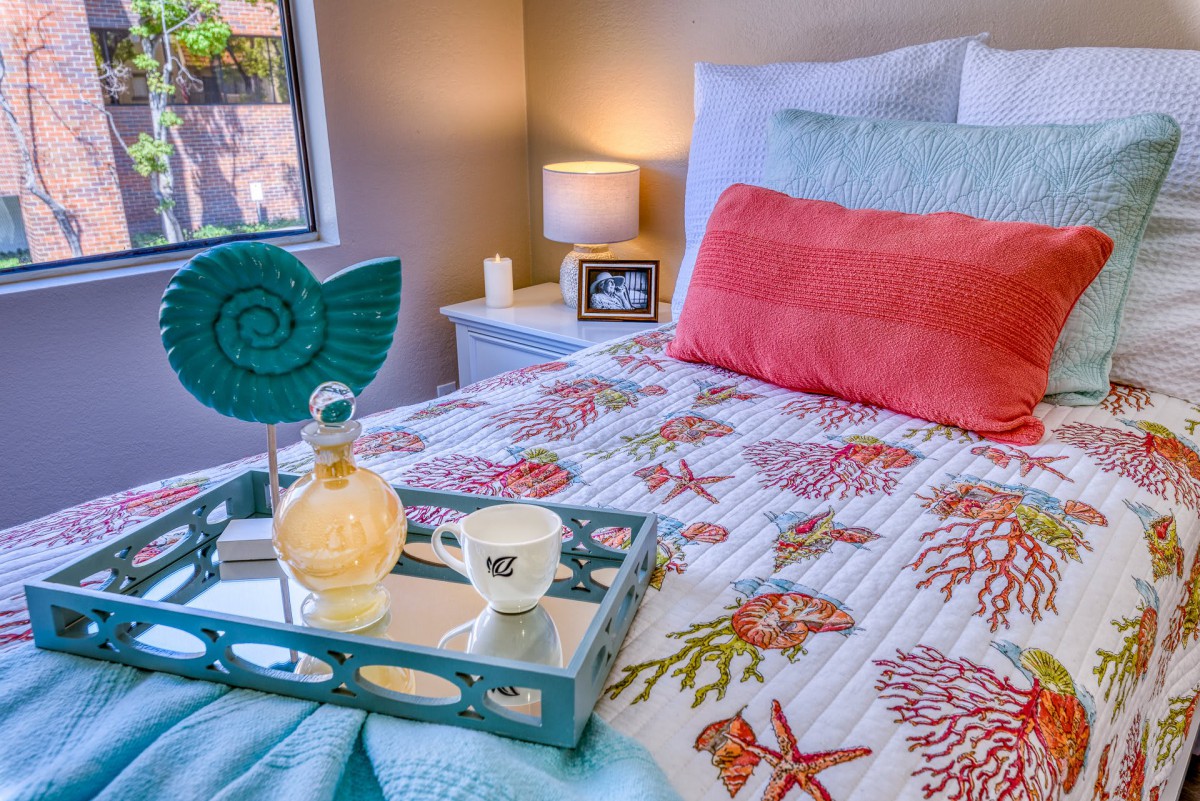 A cozy bedroom with coastal-themed bedding and decor, featuring a small lamp and a tray with a drink on the bed.