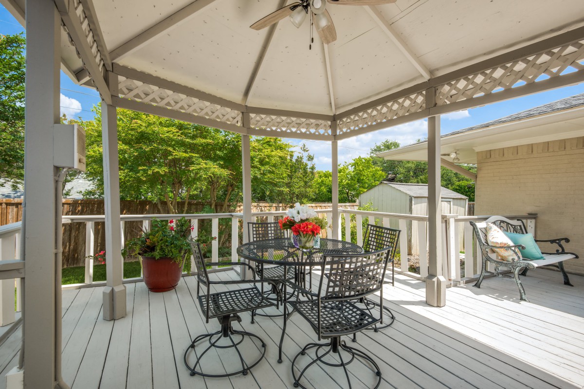 A cozy outdoor patio with a gazebo, featuring metal chairs and a table, surrounded by greenery.