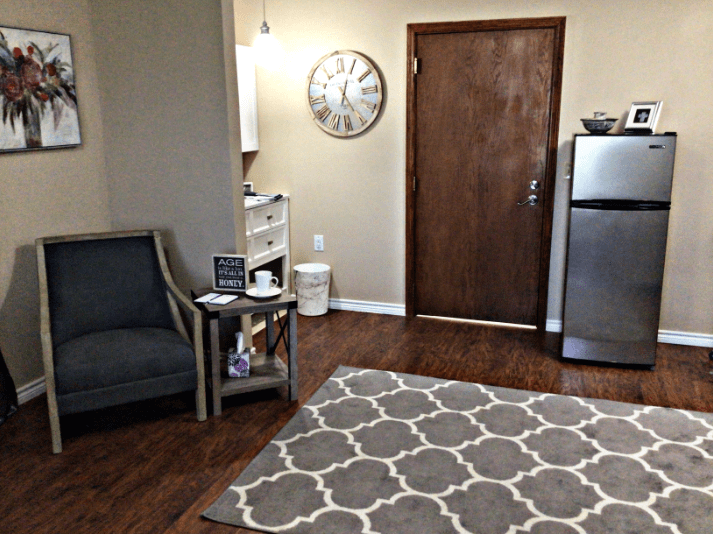 A cozy living room with a patterned rug, chair, wooden door, and a small fridge.