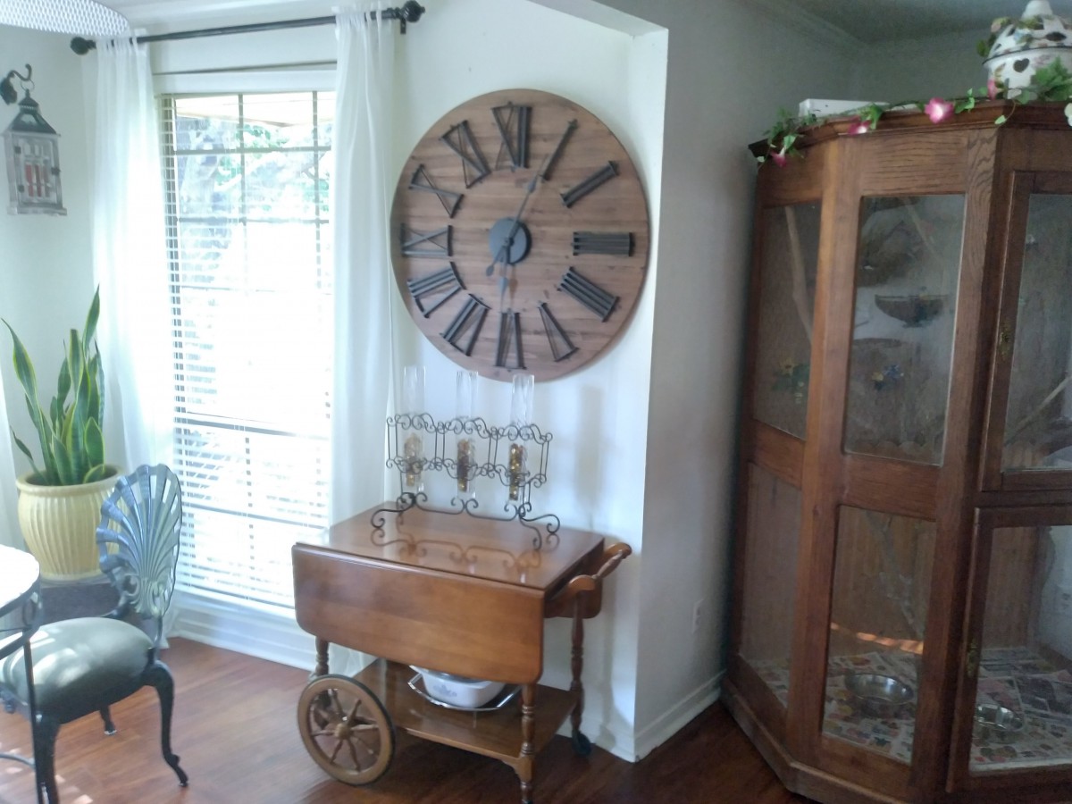 A cozy corner featuring a large wall clock, a wooden cart, and a glass cabinet beside a window.