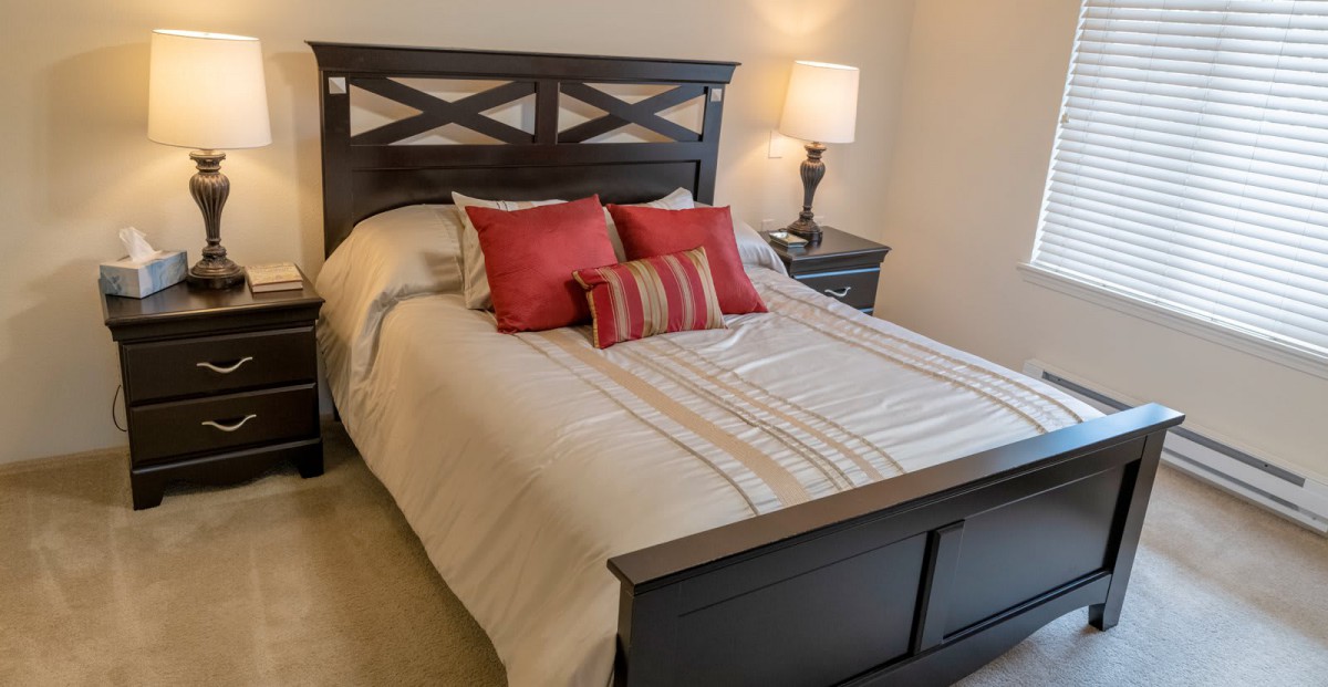 A cozy bedroom featuring a neatly made bed with red accent pillows, a wooden headboard, and a bedside lamp.