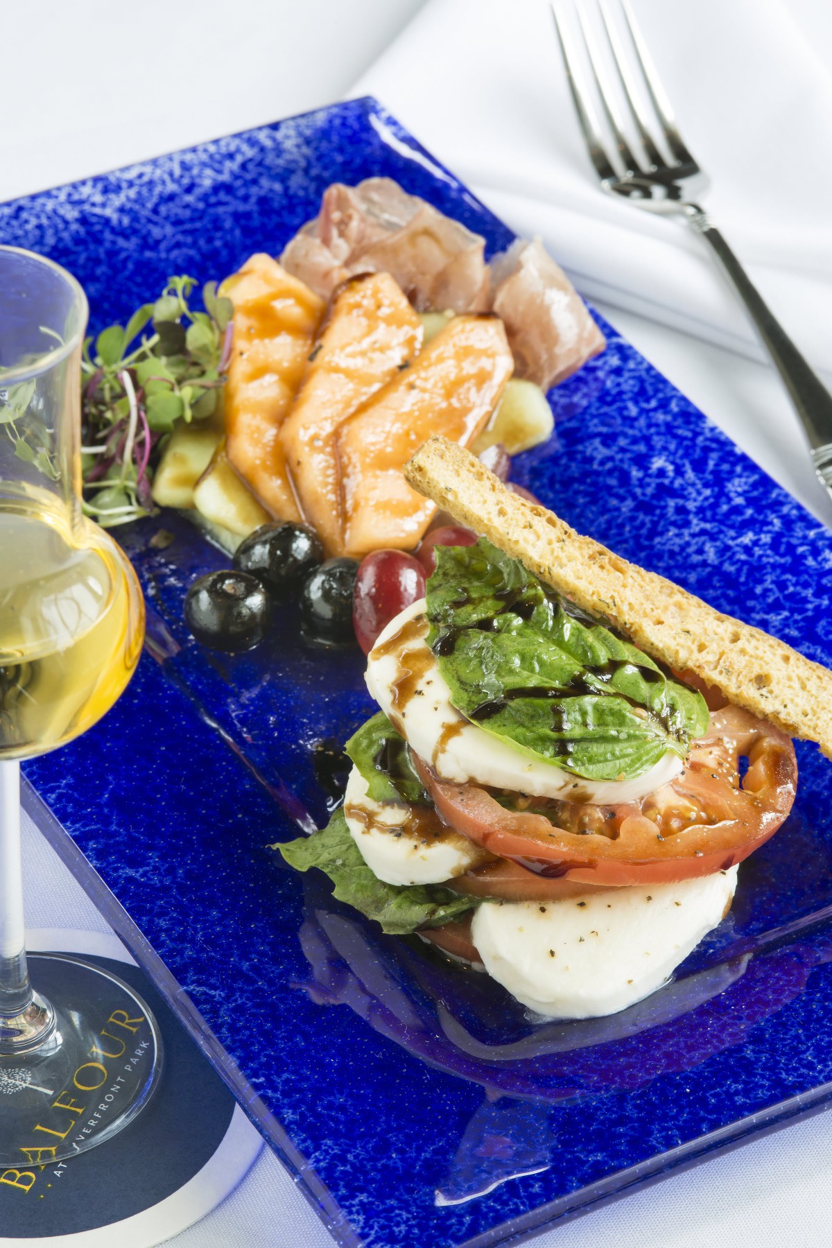 A gourmet meal featuring a Caprese salad with fresh mozzarella, tomatoes, and basil next to grilled salmon, olives, and a glass of white wine on a blue plate.