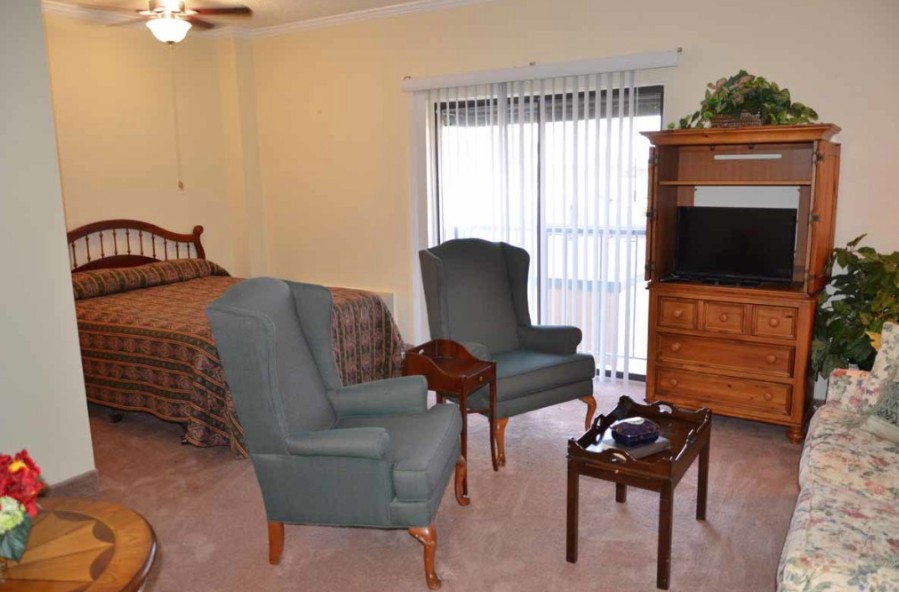 A cozy bedroom with chairs, a bed, and a TV stand near a window.