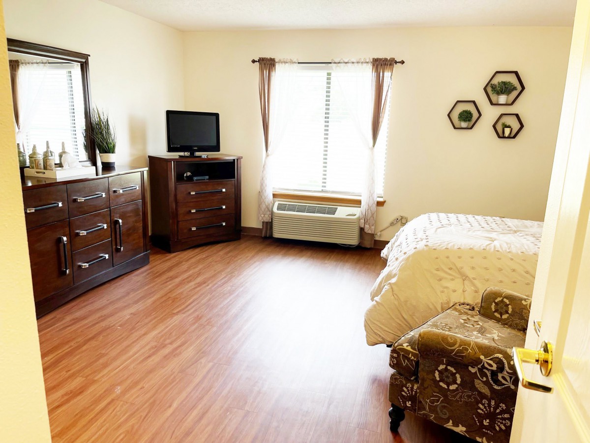 A cozy bedroom with wood flooring, featuring a bed, dresser, television, and window.