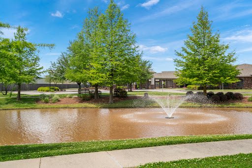 A serene park scene featuring a pond with a water fountain surrounded by lush greenery and clear blue skies.