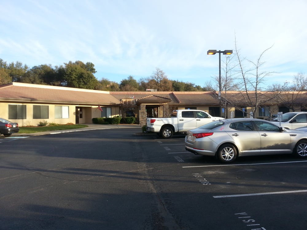 A small parking lot with parked cars in front of a single-story building, surrounded by trees during the daytime.