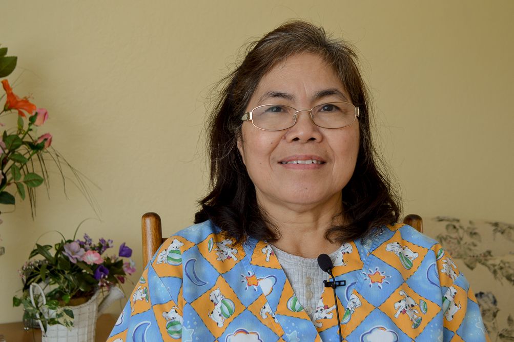 A woman with glasses smiling indoors, sitting in front of a floral arrangement.