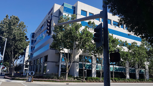 A modern office building featuring blue accents, surrounded by trees and a traffic signal.