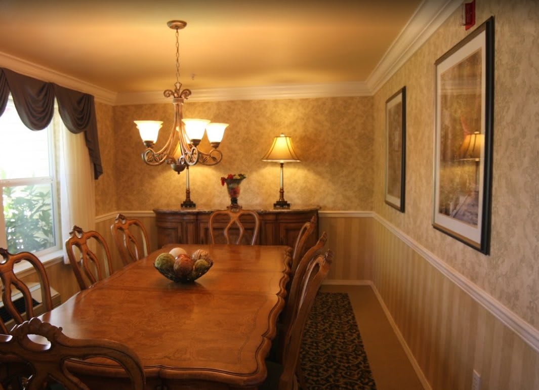 A warmly lit dining room with a wooden table and elegant chairs, featuring a chandelier and wall art.