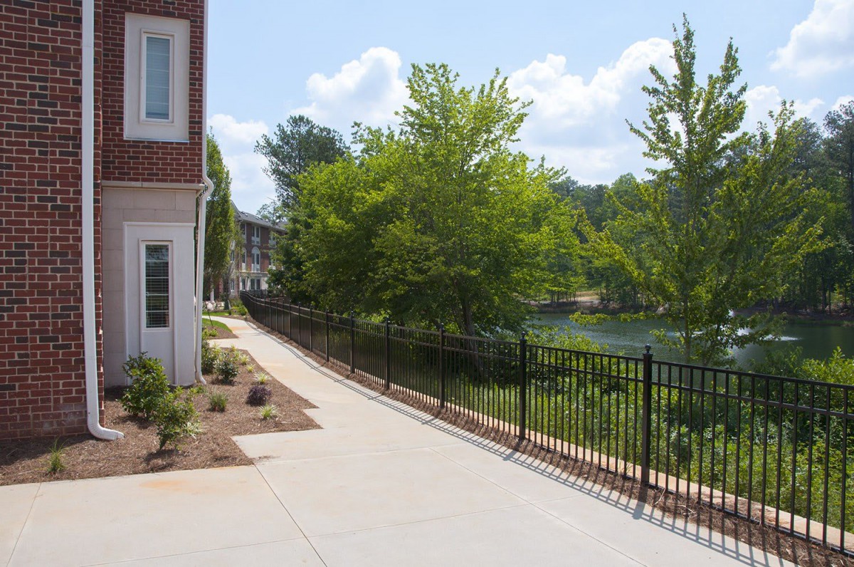 A scenic sidewalk lined with trees and a metal fence, adjacent to a lake.