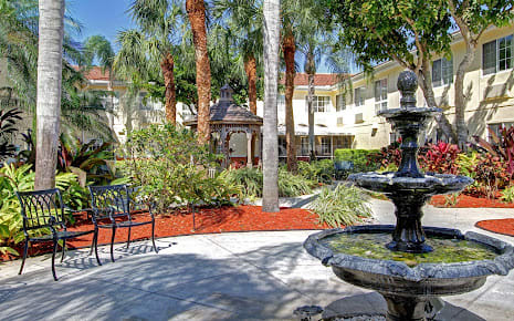 A serene garden setting with a fountain, surrounded by palm trees and outdoor seating.