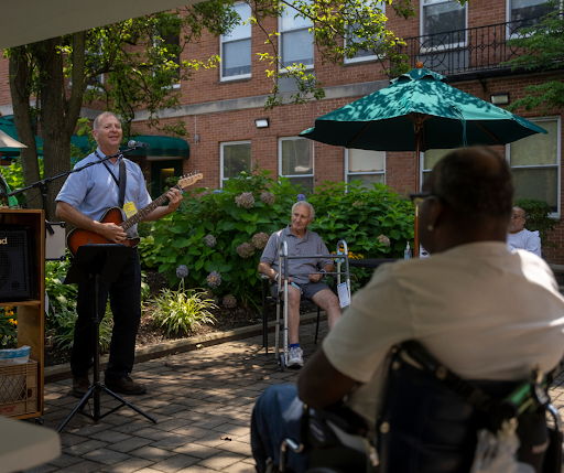 A guitarist performs for an audience of seniors enjoying a sunny day outdoors.