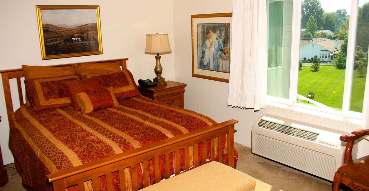A cozy bedroom featuring a wooden bed with patterned bedding, a bedside lamp, framed artwork on the wall, and white curtains covering the window.