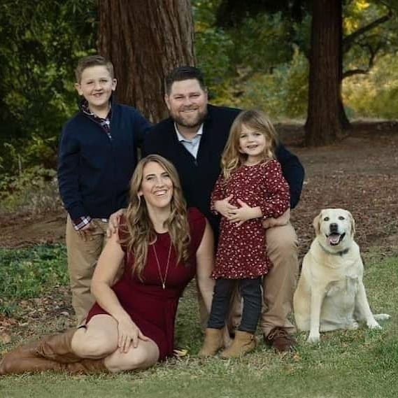A family portrait taken outdoors with parents, two children, and a dog surrounded by trees.
