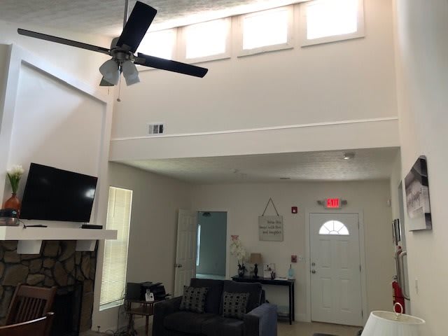 A modern living room with high ceilings and a ceiling fan, featuring a fireplace, television, and sofa. Natural light filters in through multiple small windows above.