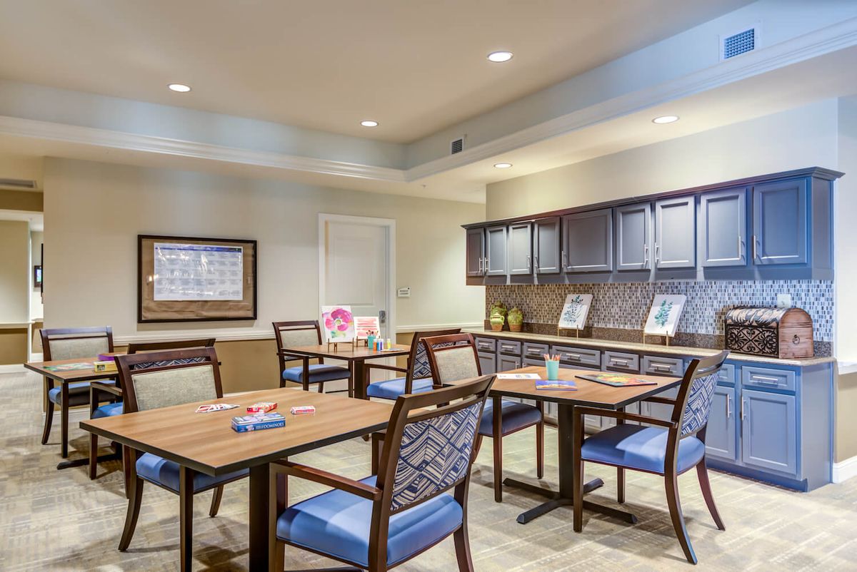 A modern dining area with wooden tables, cushioned chairs, and a small kitchenette in the background.