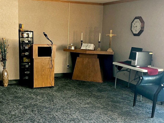 A room with a podium and microphone, an altar with candles, a clock on the wall, and a table with electronic equipment.