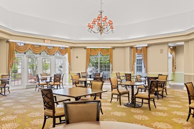 A spacious dining room with elegant decor, featuring several tables and chairs, a chandelier, and large windows allowing natural light in.