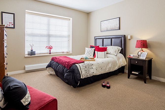 A cozy bedroom with a neatly made bed, decorated with red and white accents, next to a large window.