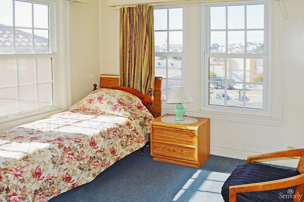 A cozy bedroom with a single bed, floral bedding, and a nightstand by large windows.