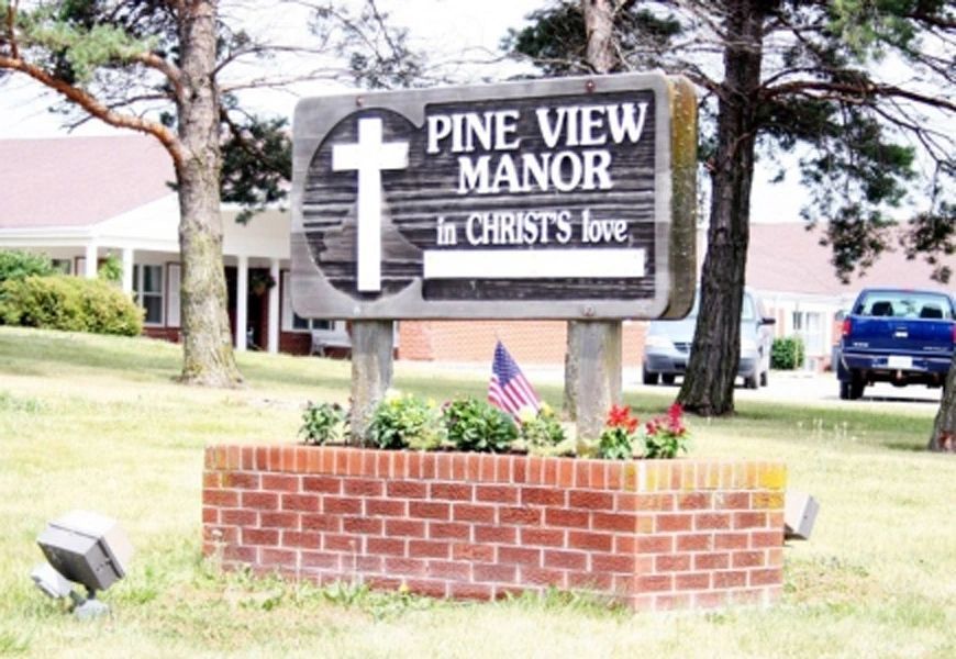A sign for Pine View Manor featuring a cross, surrounded by a brick base and an American flag.
