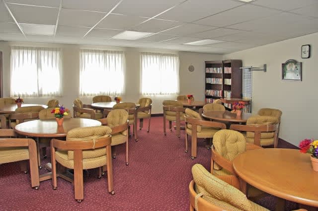 A quiet conference room with multiple round tables and cushioned chairs, featuring carpet flooring and natural light from several windows.