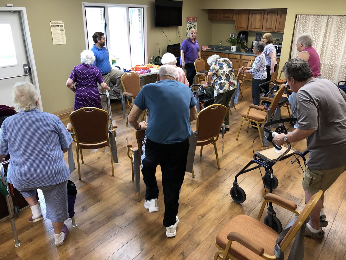 A group of elderly individuals participating in an exercise class indoors, using chairs for support.