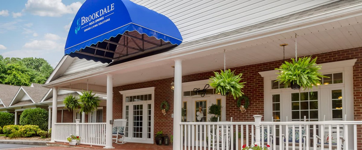 A building entrance featuring a blue awning, porch with columns, hanging plants, and white railings.