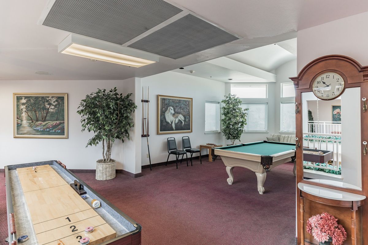 A spacious game room featuring a pool table and shuffleboard, complemented by large windows and decorative plants.