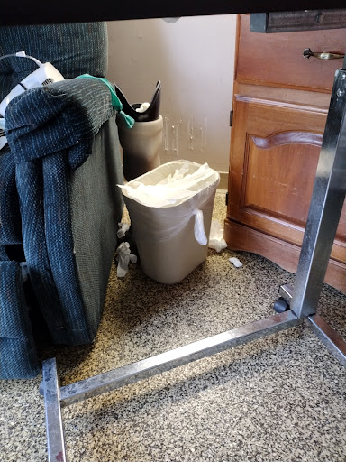 A cluttered corner showing a trash can next to some furniture and a wooden cabinet.