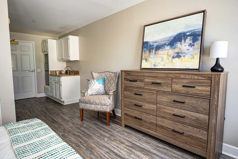 A modern bedroom with a wooden dresser, patterned armchair, and framed artwork on the wall.