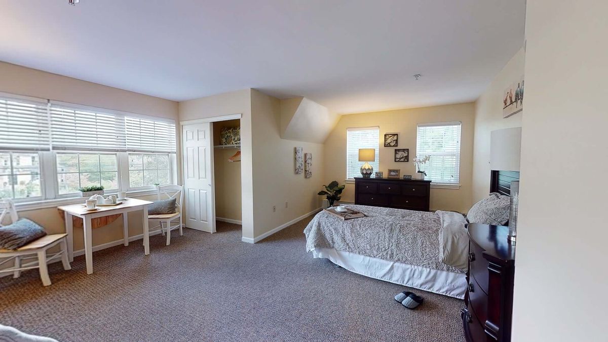 A spacious bedroom with a neatly made bed, dresser, and natural light streaming through large windows.