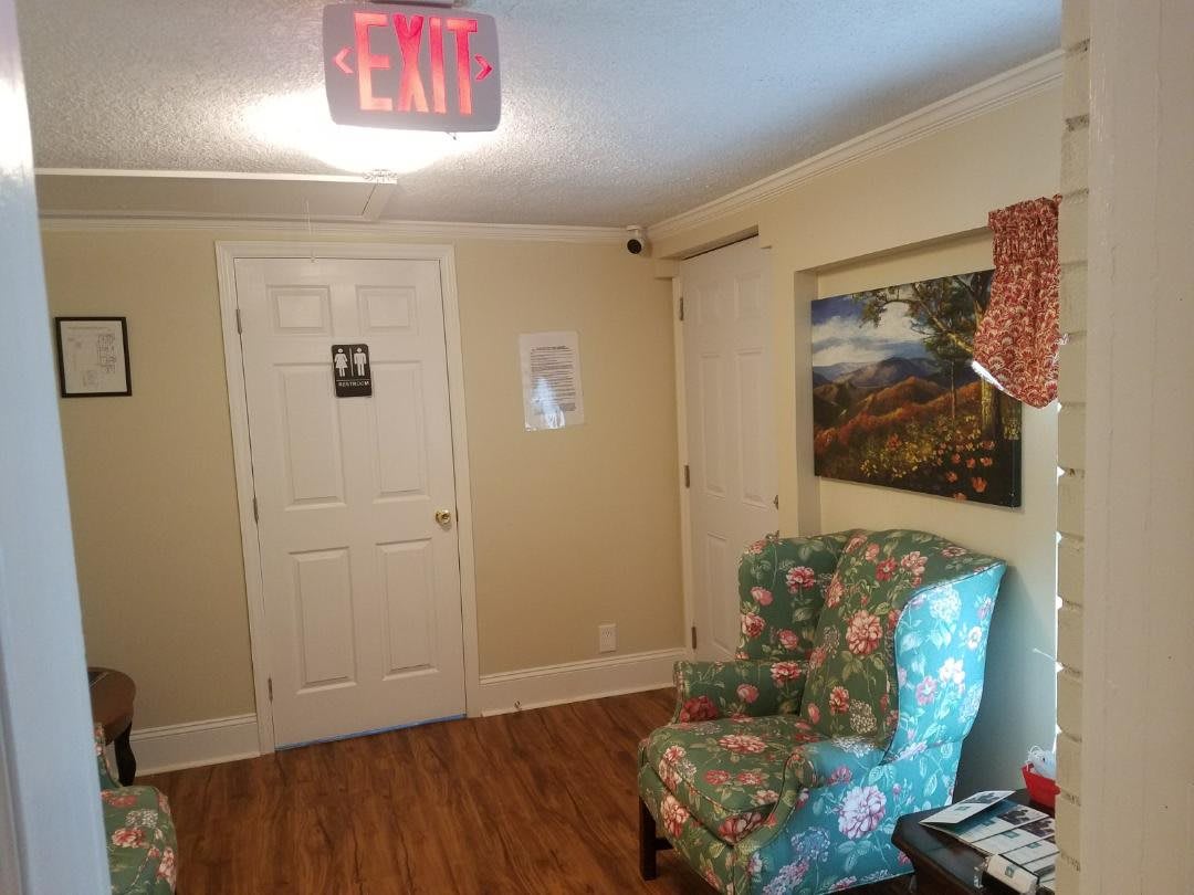 A cozy waiting room interior with floral armchairs, a doorway, and an illuminated exit sign.