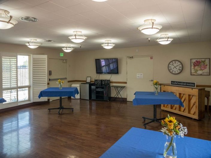 A well-lit room with tables set with blue tablecloths, a wall-mounted television, flowers in a vase, and a clock on the wall.