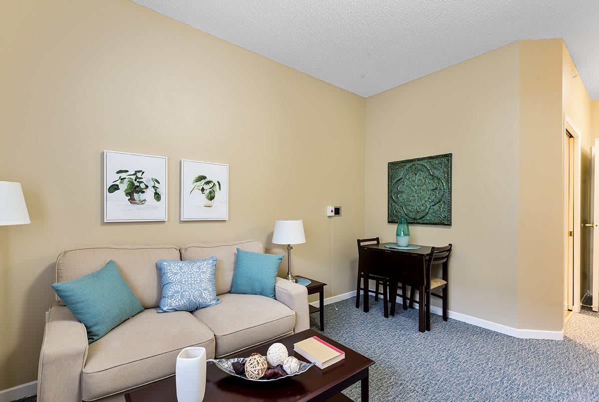 A cozy living room with a beige sofa, decorative pillows, and a coffee table, accented by wall art and a small dining area.