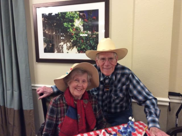 An elderly couple wearing plaid shirts and hats, smiling while seated indoors with a checkered tablecloth and a picture frame in the background.