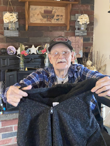 An elderly man smiles while holding a jacket indoors.
